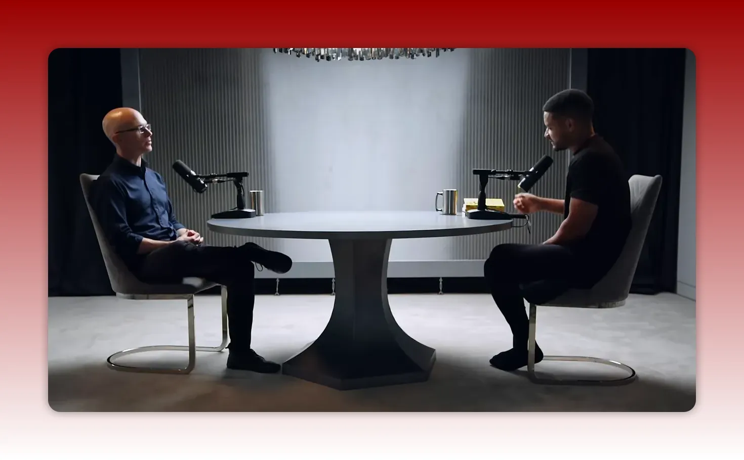 Wide studio shot of two people seated across a round table with microphones, one gesturing as they speak; neutral backdrop.