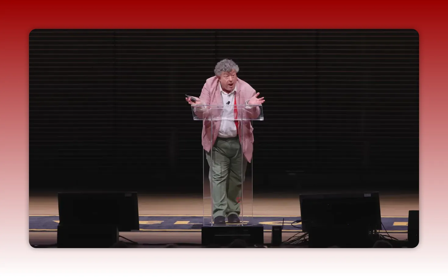 Full-stage view of speaker with arms outstretched behind a clear lectern, expressing surprise