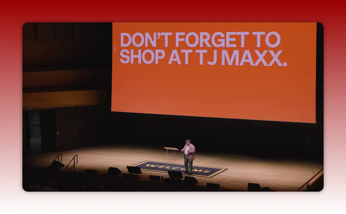Wide shot of a presenter on stage beneath a large orange slide, showing an auditorium setting