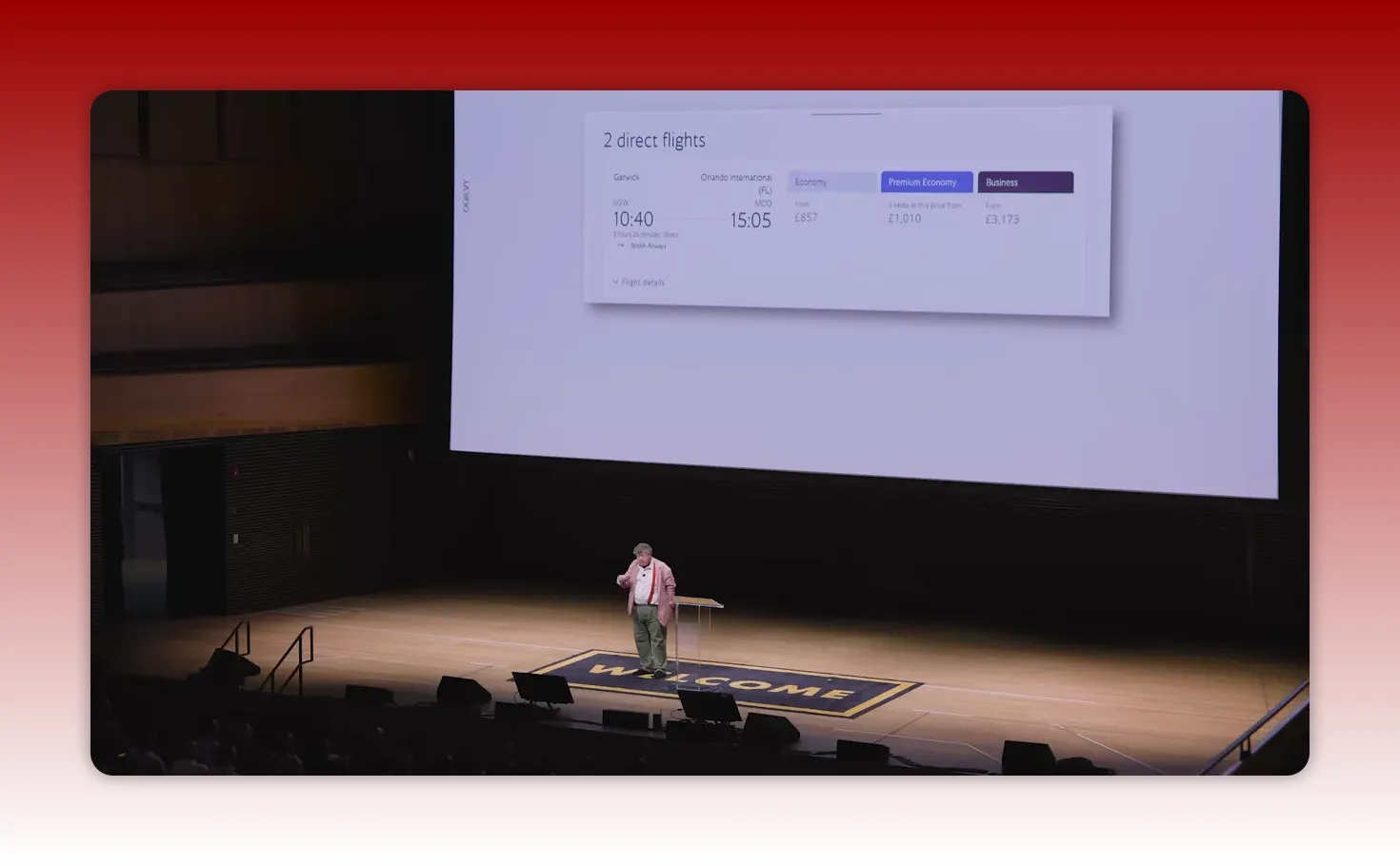 Wide auditorium shot with presenter and a large projected slide showing flight pricing tiers (economy, premium economy, business).