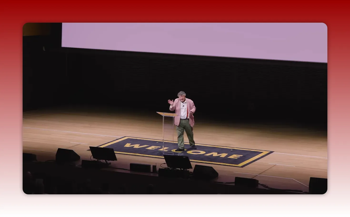 Wide, high-quality shot of the speaker at a clear podium standing on a 'WELCOME' mat at centre stage, with clear lighting and composition.