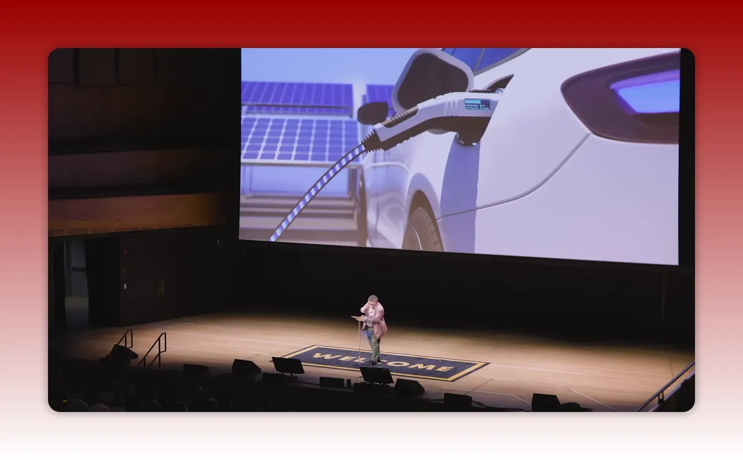Large presentation slide showing an electric vehicle being charged with the speaker on stage below