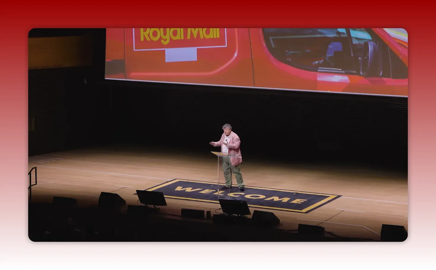 Speaker on stage with a large screen showing a red Royal Mail van; 'WELCOME' mat visible on stage