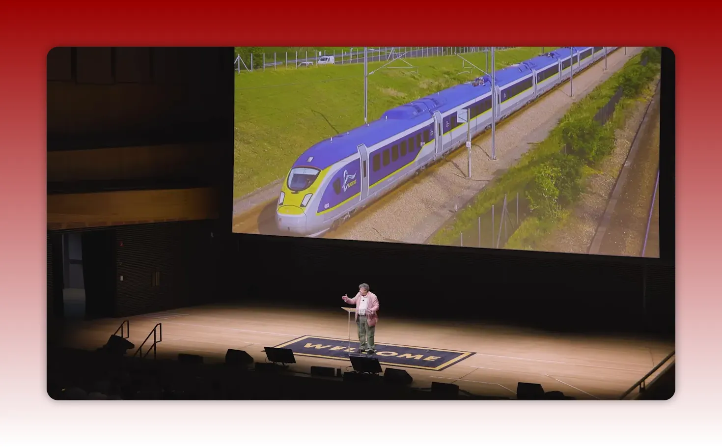 Wide shot of a presenter on stage with a large projected image of a blue and yellow train, showing the train example.