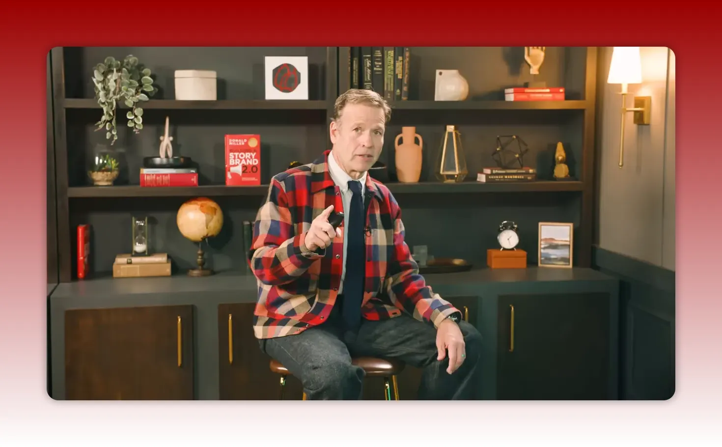 Presenter seated on a stool in front of a bookshelf-backed studio set, pointing toward the camera as he introduces examples.