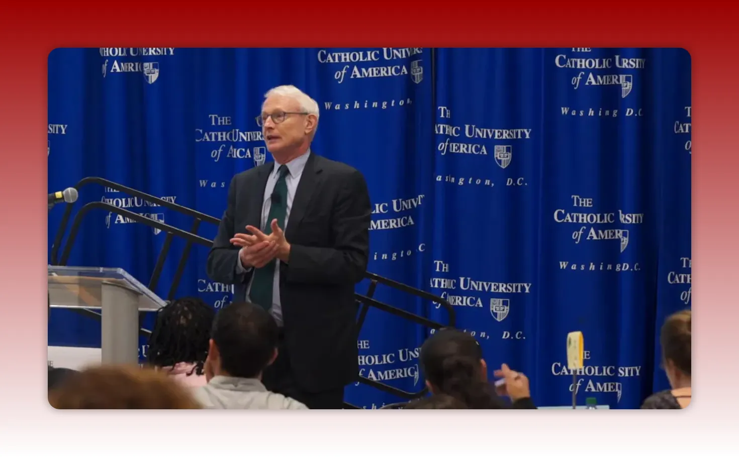 speaker gesturing beside a podium with audience in foreground and university-branded curtain behind