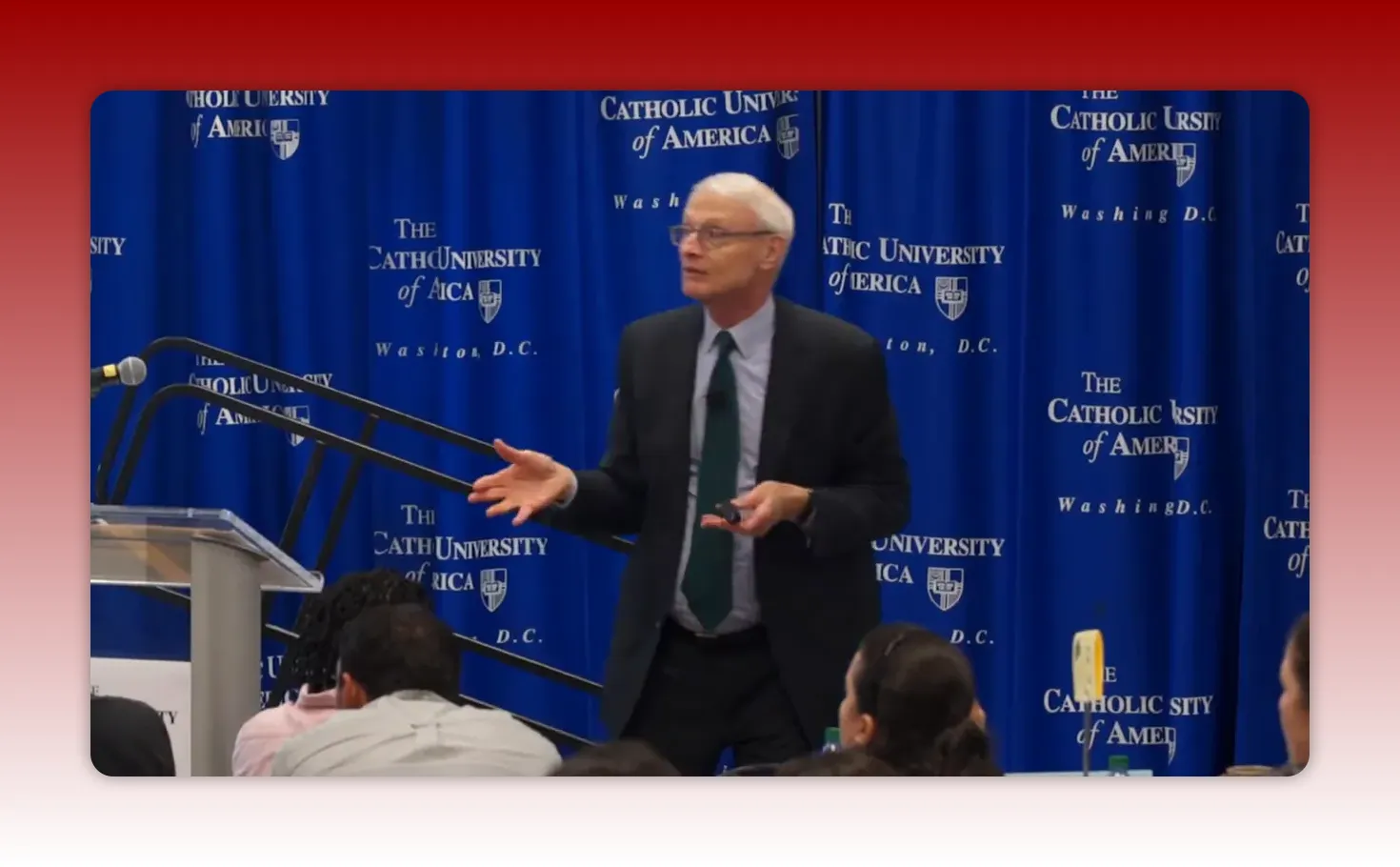 Presenter standing in front of a blue university backdrop, hands open while explaining industry analysis to an audience.