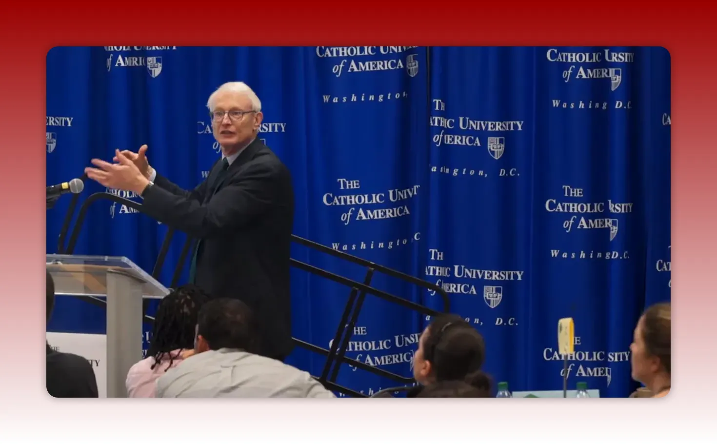 presenter facing the audience and gesturing with open hands in front of a blue 'The Catholic University of America' backdrop