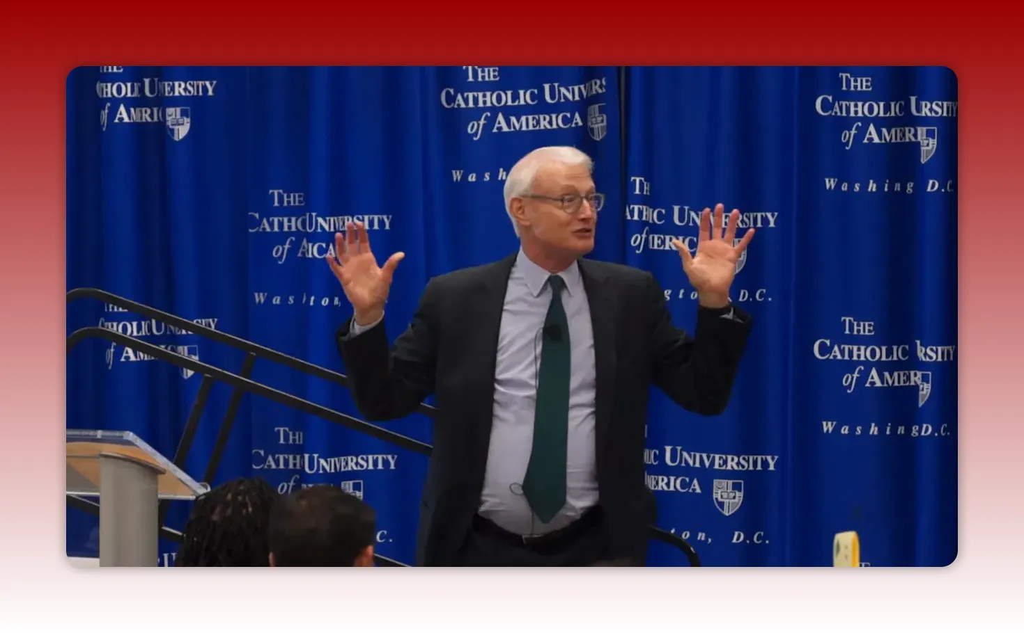 Presenter on stage gesturing with both hands in front of a blue university backdrop to emphasize a point.