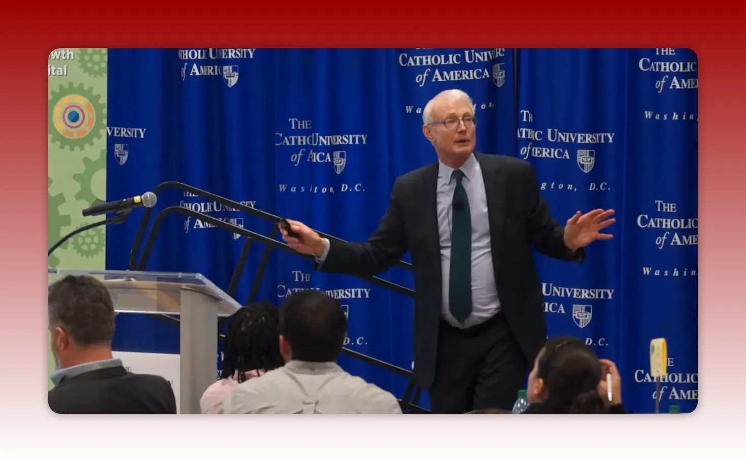 Presenter on stage gesturing with a remote in front of a university-branded backdrop, addressing the audience