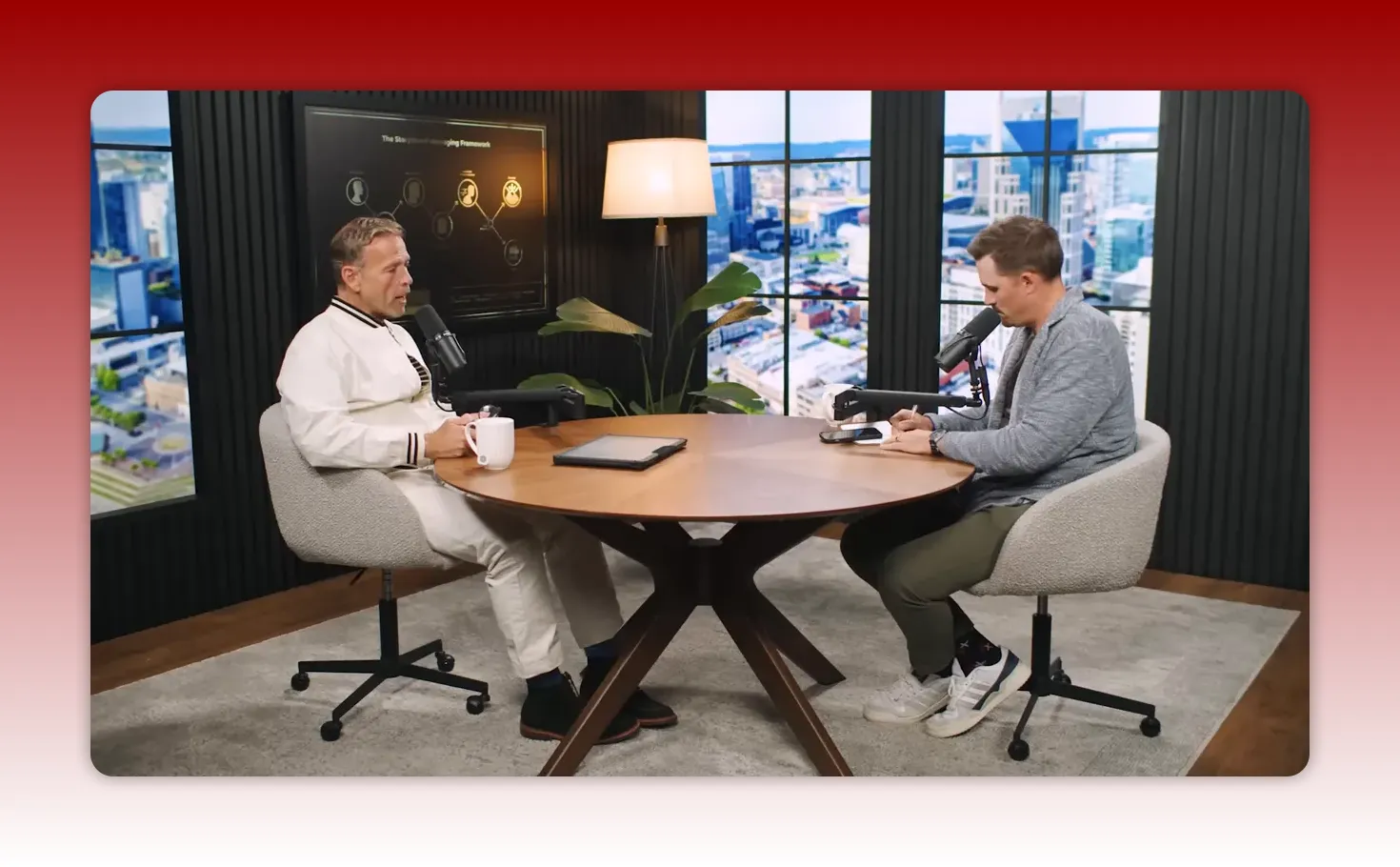 Two hosts at a round table, one speaking into a microphone while the other takes notes.