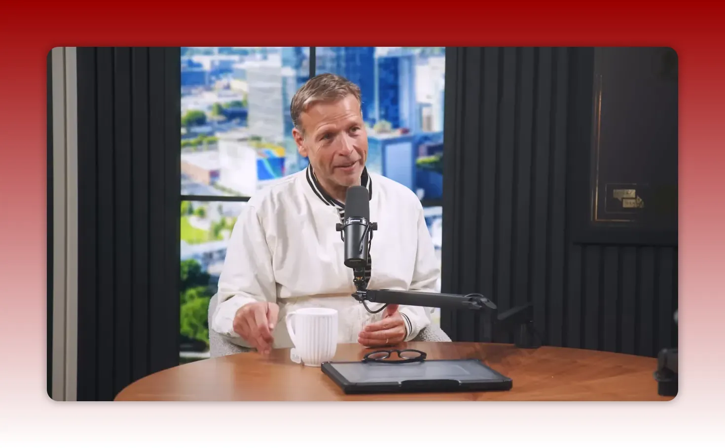 Well-lit podcast host smiling and speaking into a microphone at a wooden table with a mug and tablet in view, illustrating distribution and discipline.