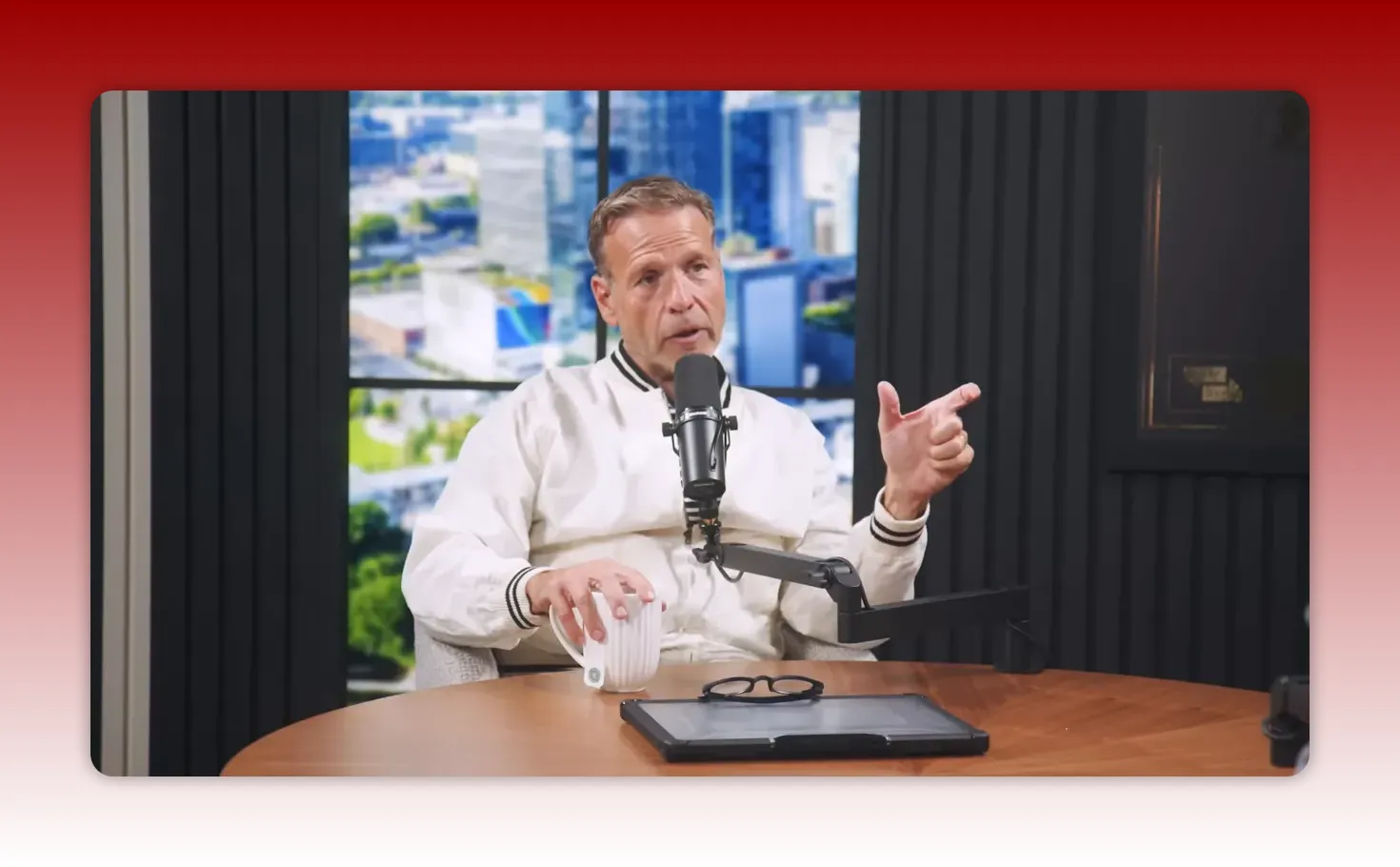 Podcast host making a directed pointing gesture while speaking into a microphone with a tablet and mug on the table.