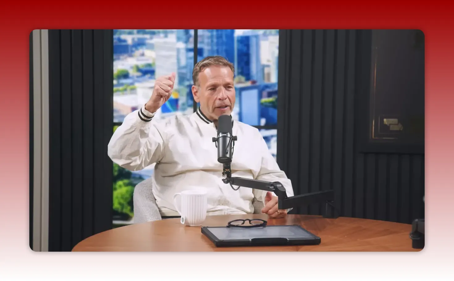 Podcast host energetically gesturing with an upward hand while speaking into a microphone; mug, glasses and tablet visible on the table suggesting a practical demo.