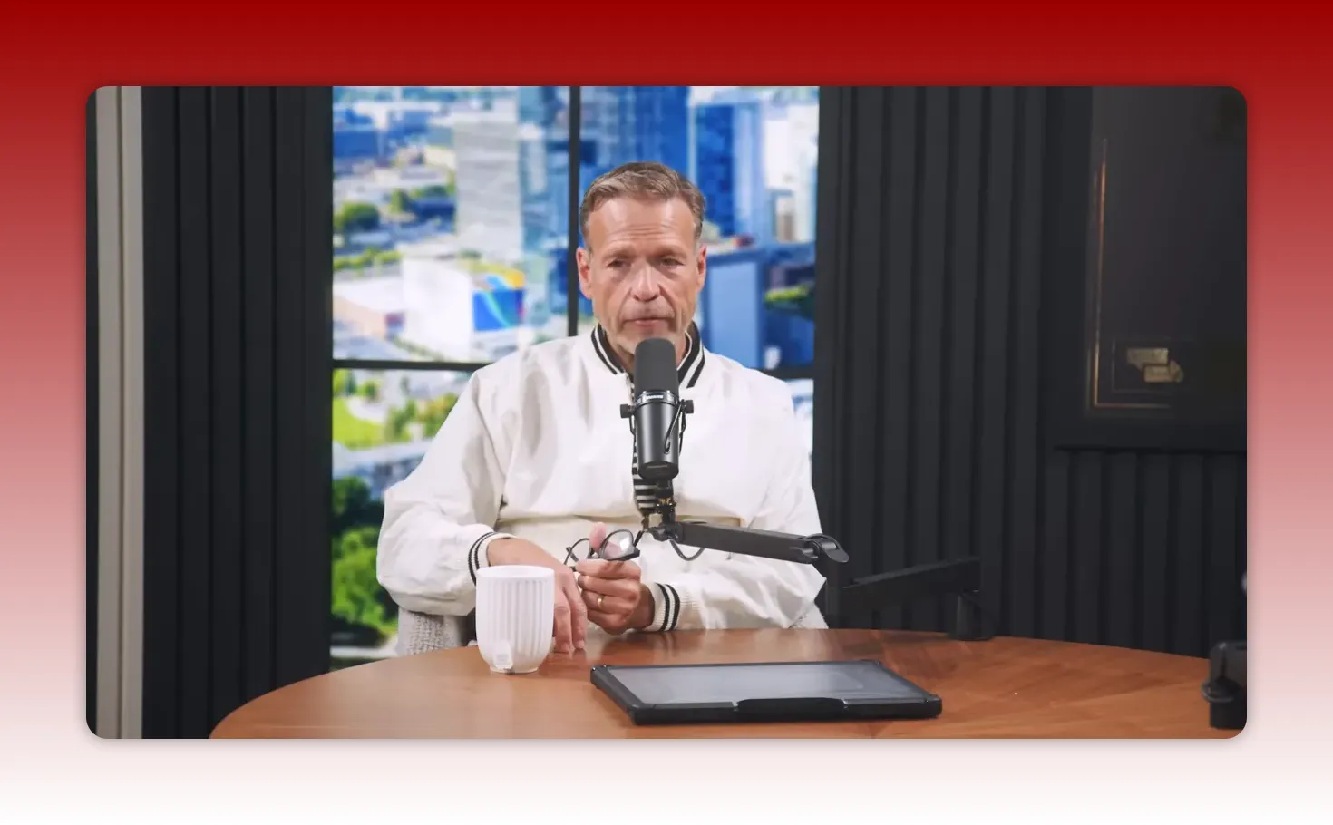 Centered podcast host speaking into a microphone with a mug and tablet on the desk.
