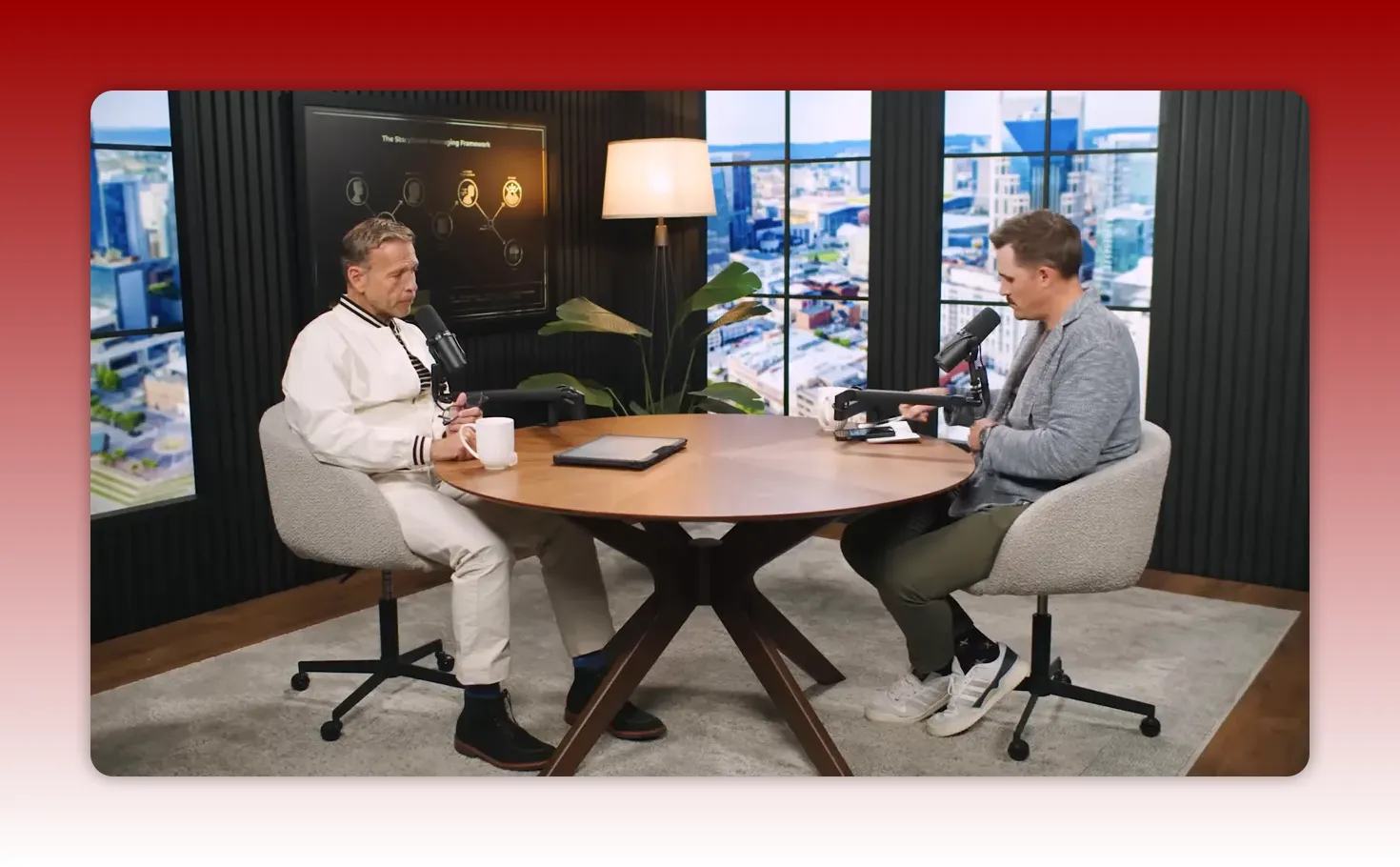 Wide studio shot of two hosts conversing at a round table with a presentation screen and city-view windows behind them.