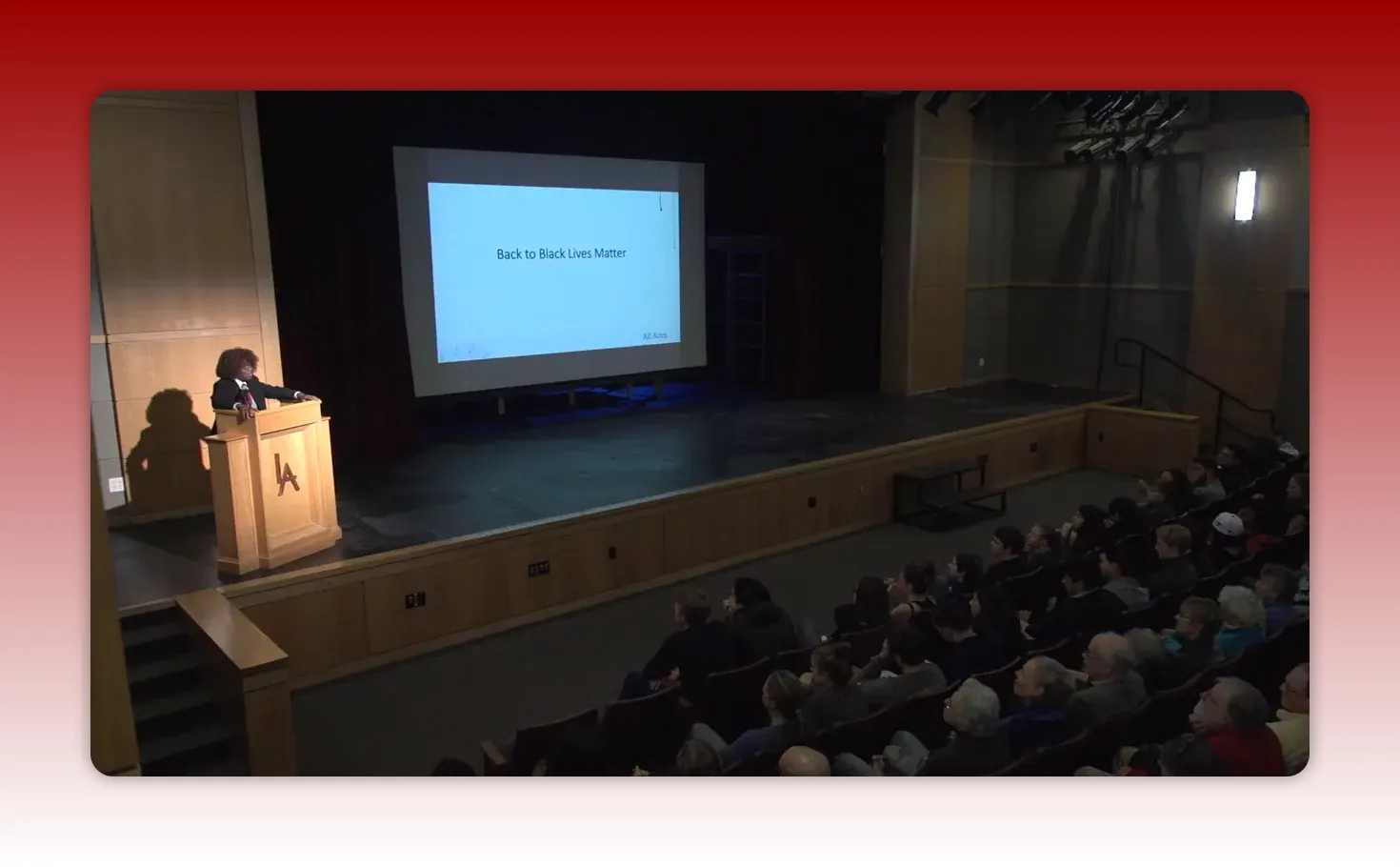 Clear wide shot of the lecture stage with the presenter at the podium and a projection screen reading 'Back to Black Lives Matter' as the audience watches.