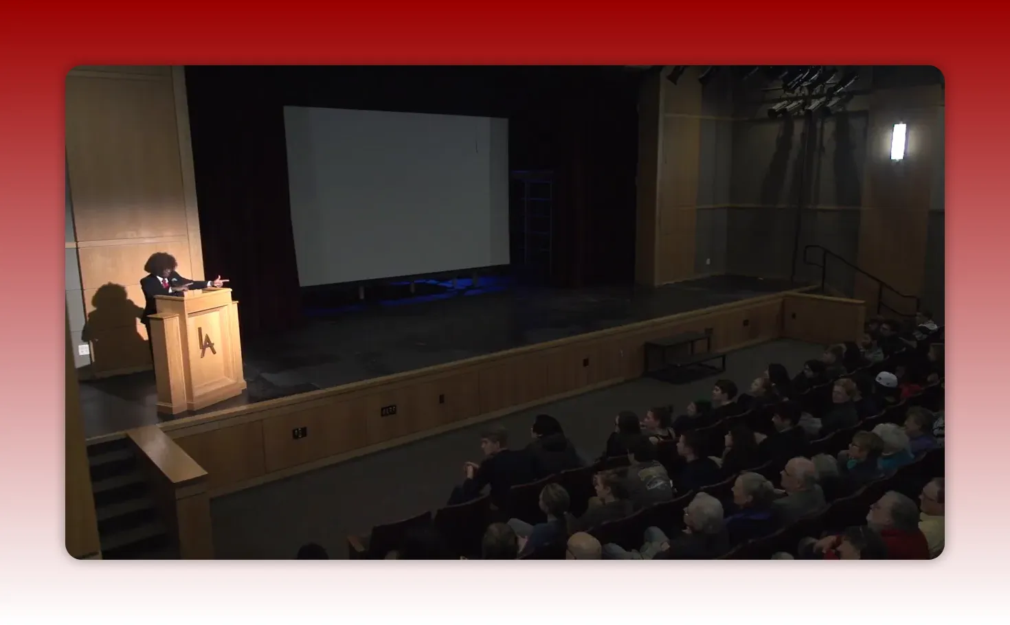 Wide auditorium shot showing the presenter at a lectern making a hand gesture with the seated audience clearly visible; suitable for illustrating concluding or reflective remarks.
