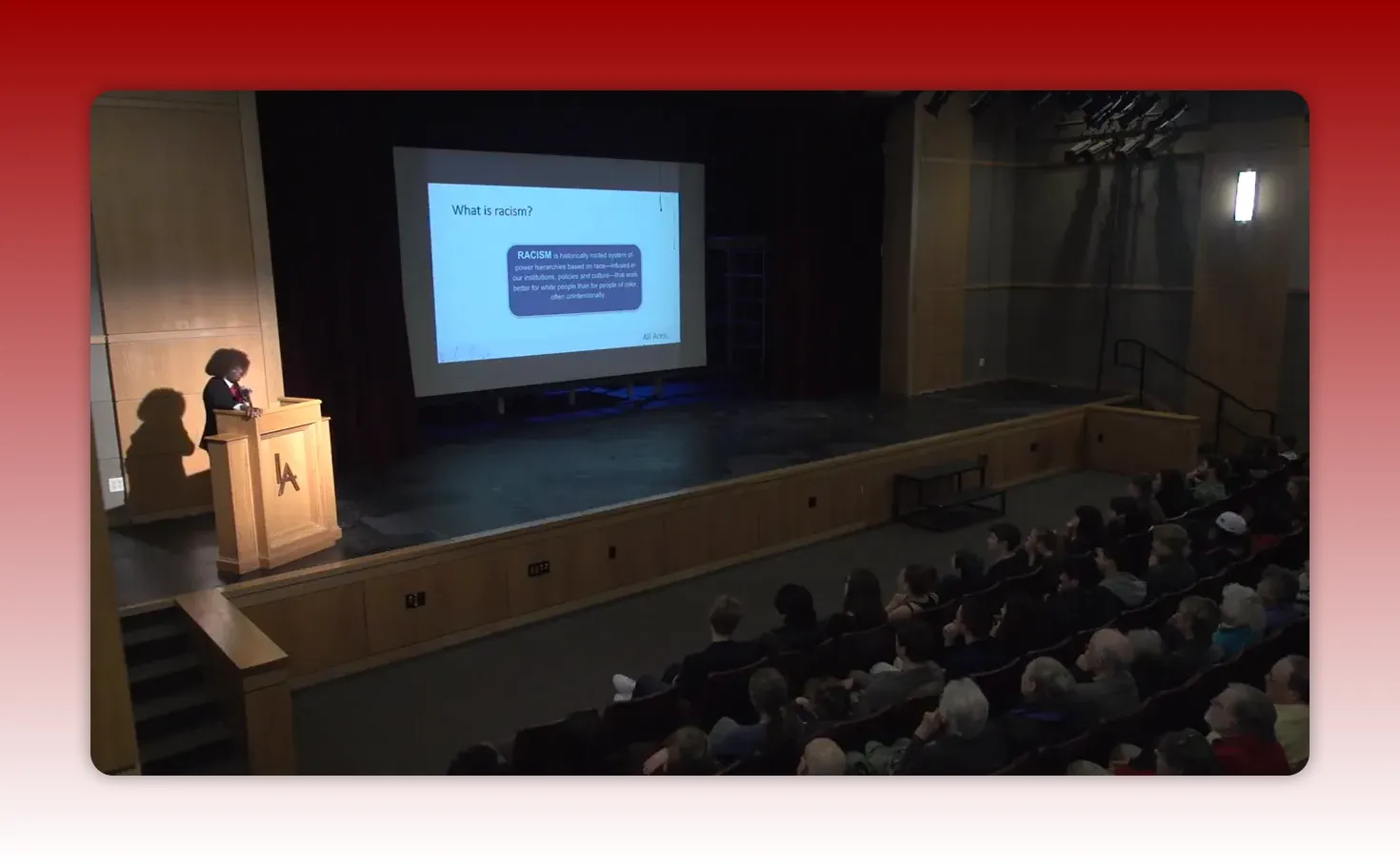 Clear auditorium shot of speaker at podium with a projected slide titled 'What is racism?'