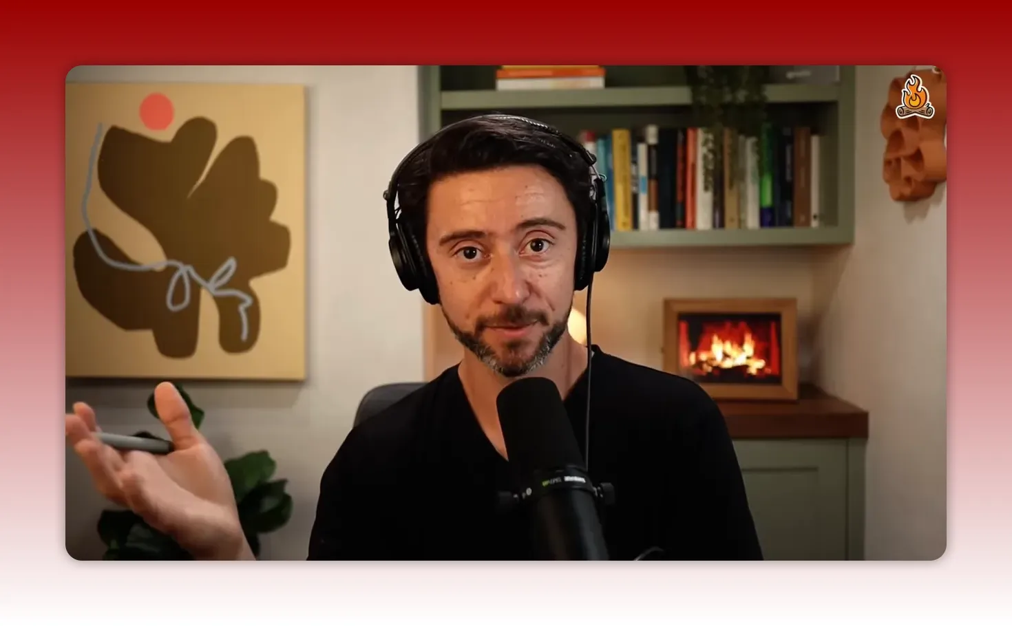 Host facing the camera wearing headphones and speaking into a microphone with a bookshelf and lit fireplace in the background.