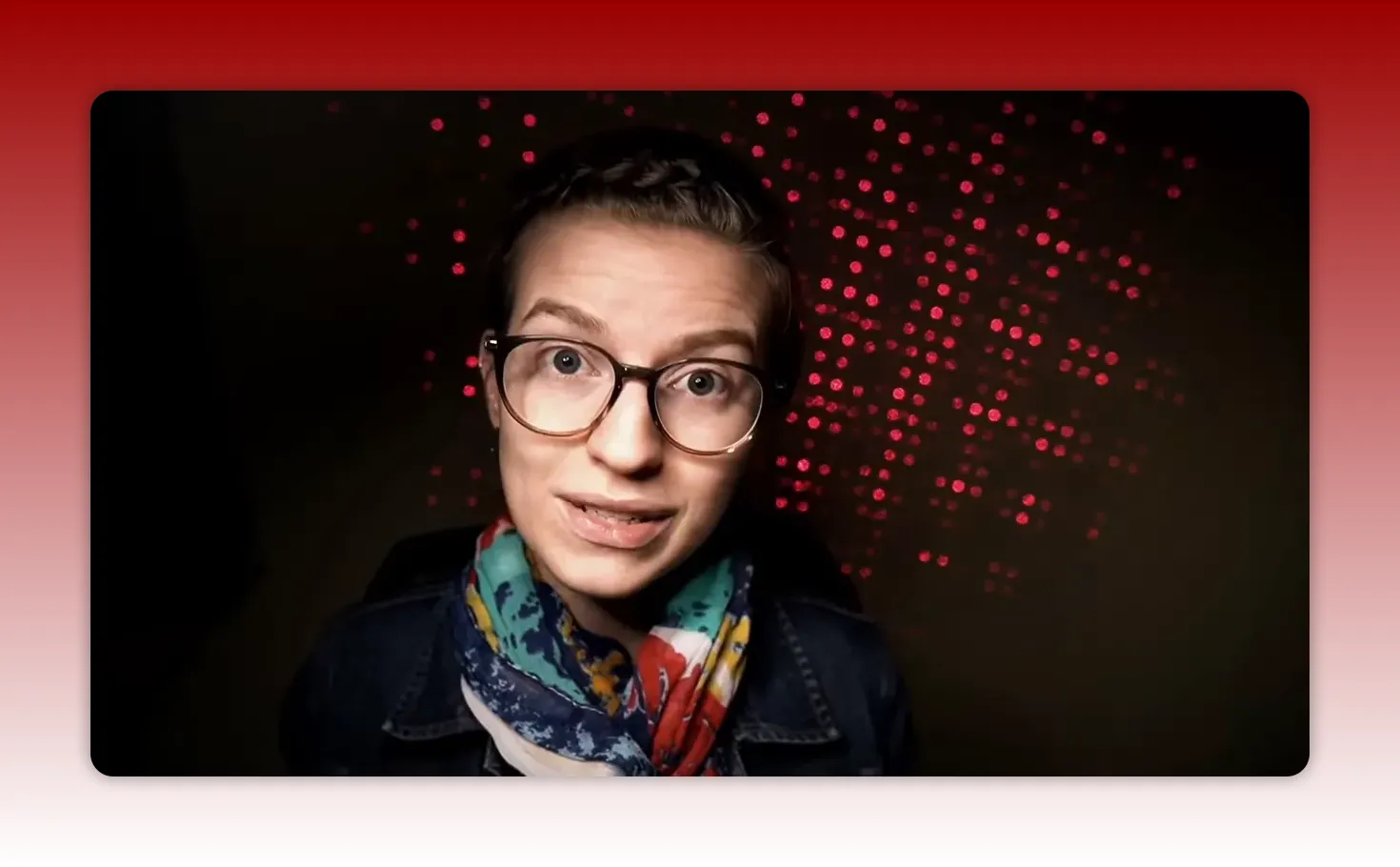Well-lit head-and-shoulders shot of a presenter looking at camera with glasses and a colorful scarf; red dotted lights form a subtle background pattern.