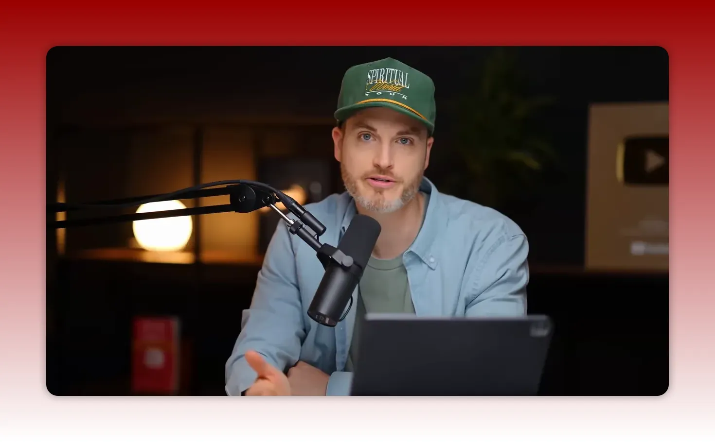 Host speaking directly to camera with a boom microphone, tablet in front, and studio YouTube play button in the background.