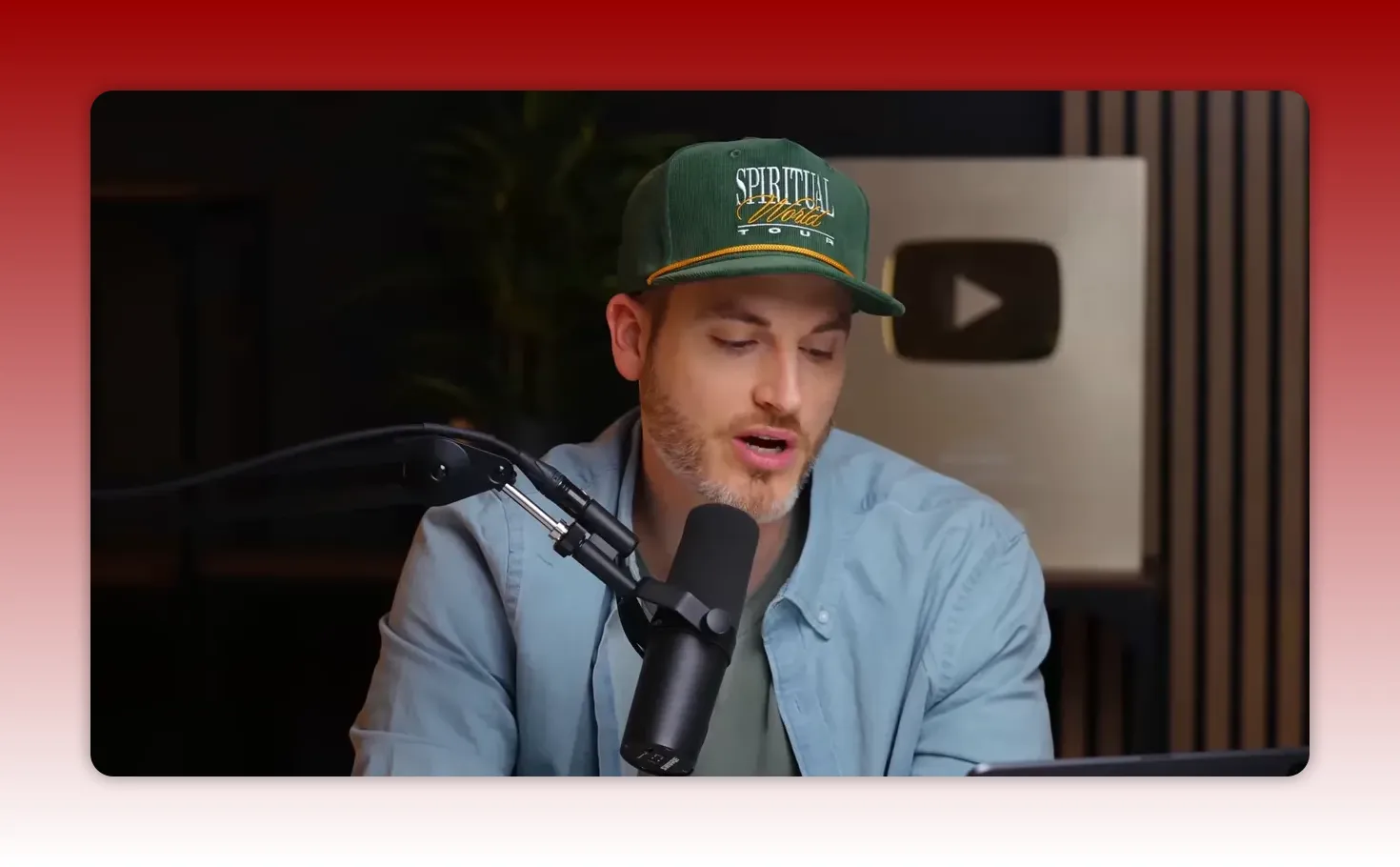 Presenter speaking into a studio microphone with a visible YouTube play button in the background, mid-sentence and clearly lit.