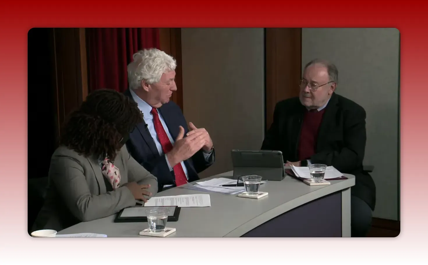 Three panelists at a table, the central panelist speaking with hand gestures while others listen, tablet and papers visible on the table.