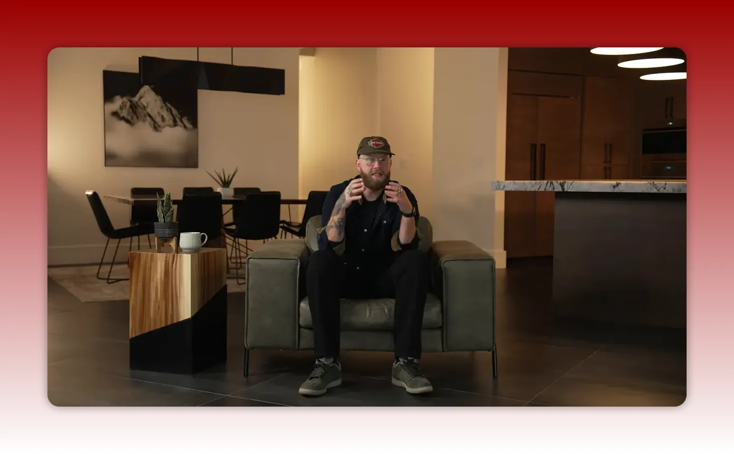 Wide, well-lit shot of the presenter seated in an armchair in a living-room/kitchen set using hand gestures