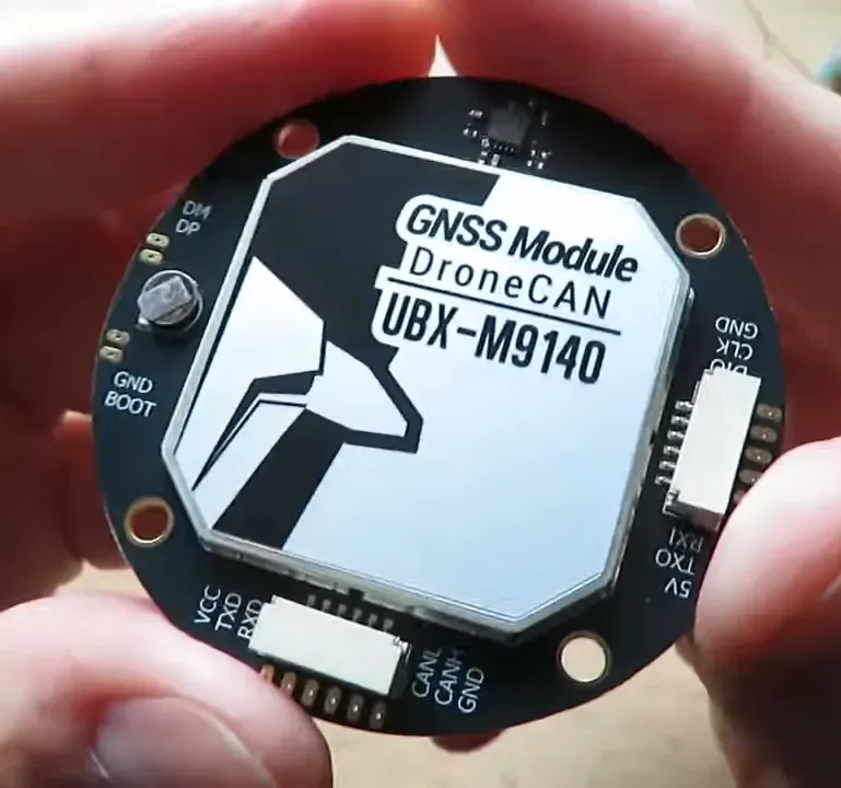 Close-up view of a round GNSS PCB held between two fingers showing the antenna cover and connector pads