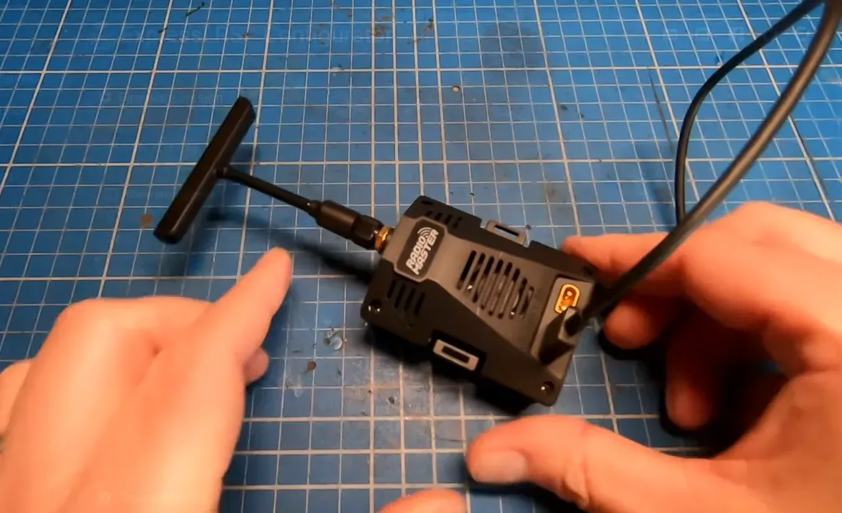 Top‑down view of a RadioMaster Ranger Micro backpack on a blue cutting mat with antenna and USB cable connected, hands nearby