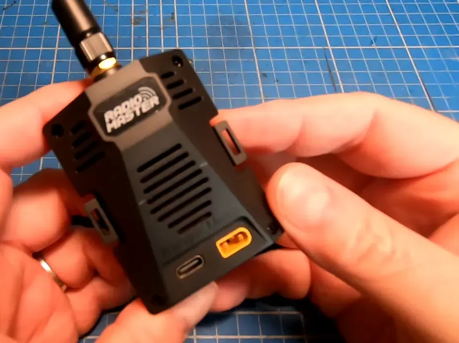 Wider view of a RadioMaster Ranger Micro backpack held in both hands on a blue cutting mat, with antenna and visible USB and power ports.