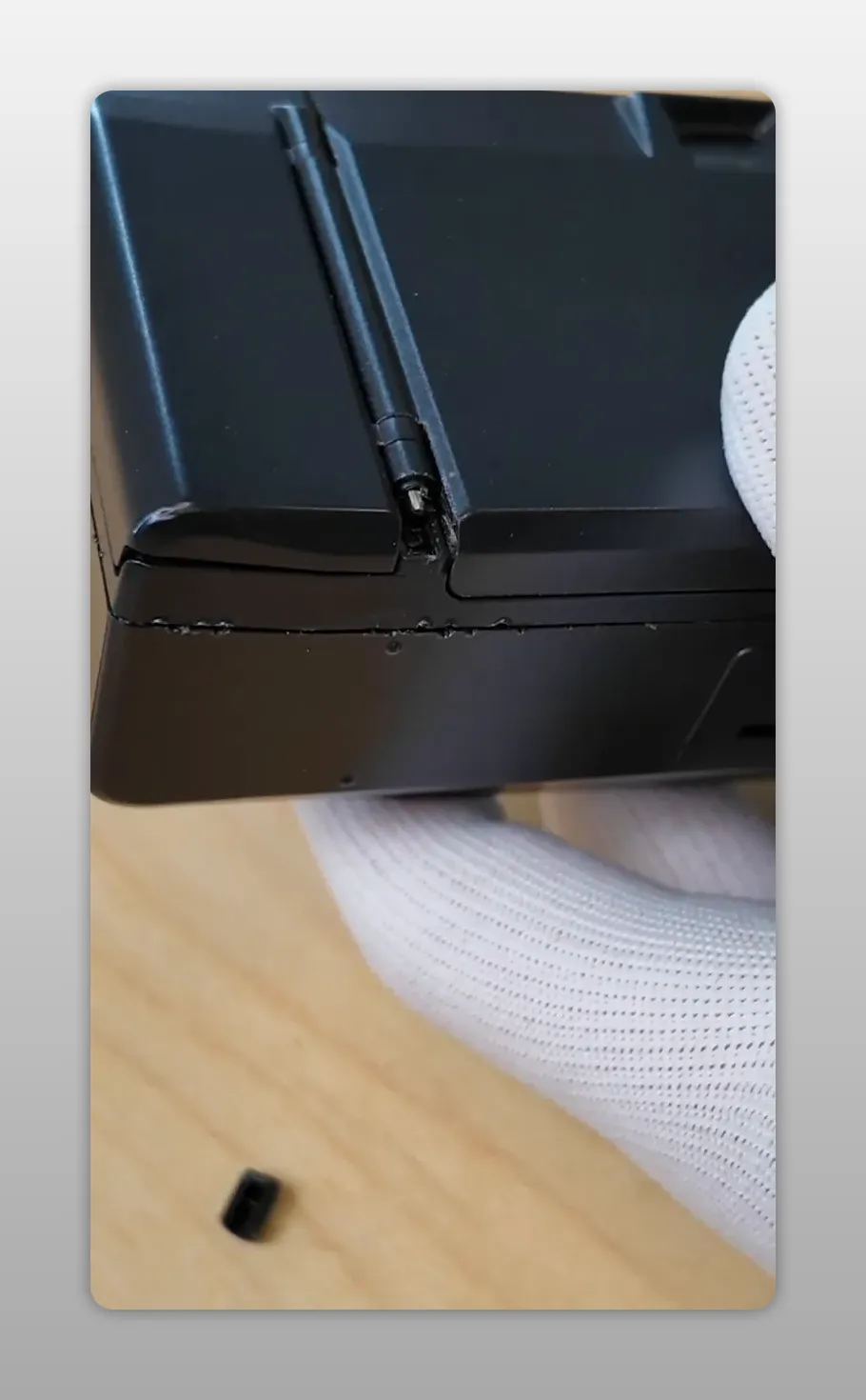 Vue claire de la charnière du Yashica T3 avec la goupille de maintien exposée et la petite protection en plastique posée sur la table, un gant tient l’appareil.