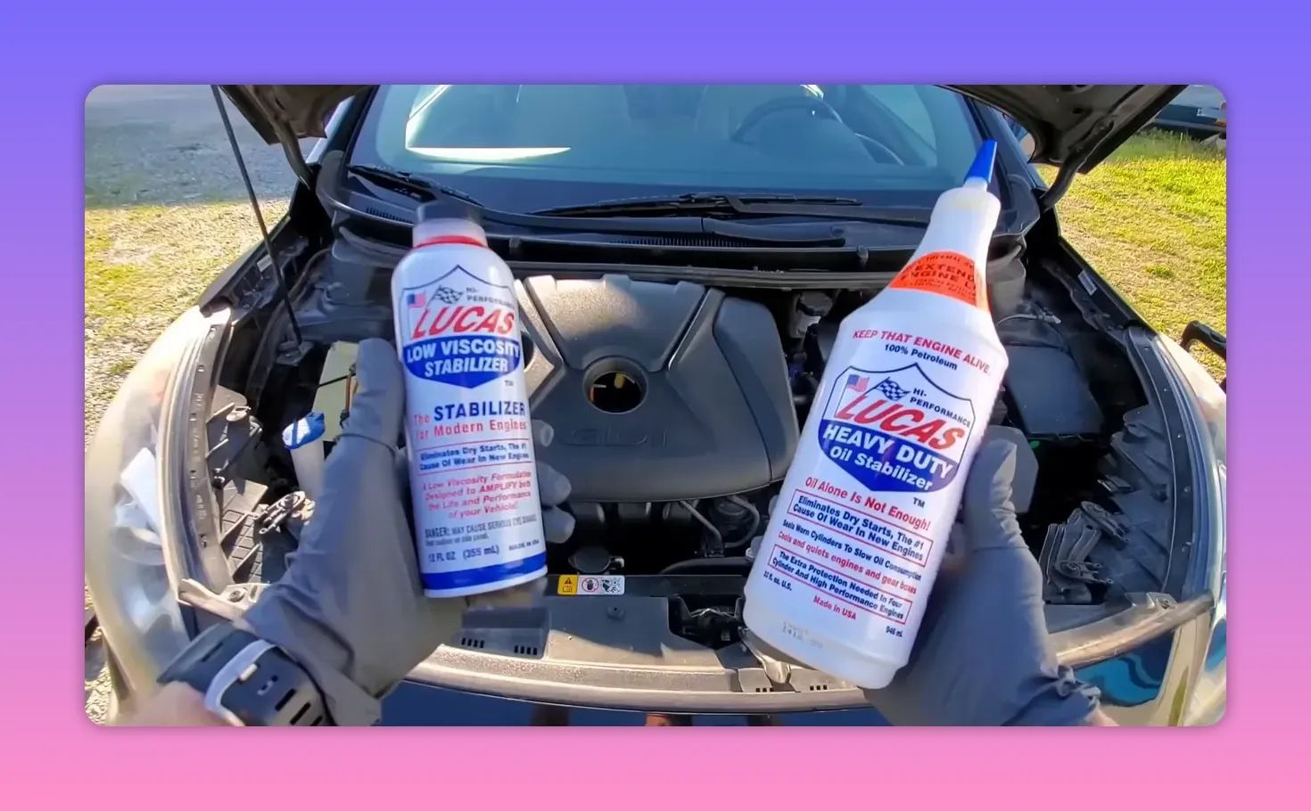 Hands holding two Lucas oil stabilizer bottles (Low Viscosity and Heavy Duty) over an open engine bay