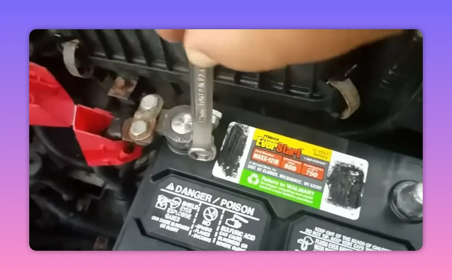 Wrench loosening a car battery terminal clamp with a red protective cover nearby