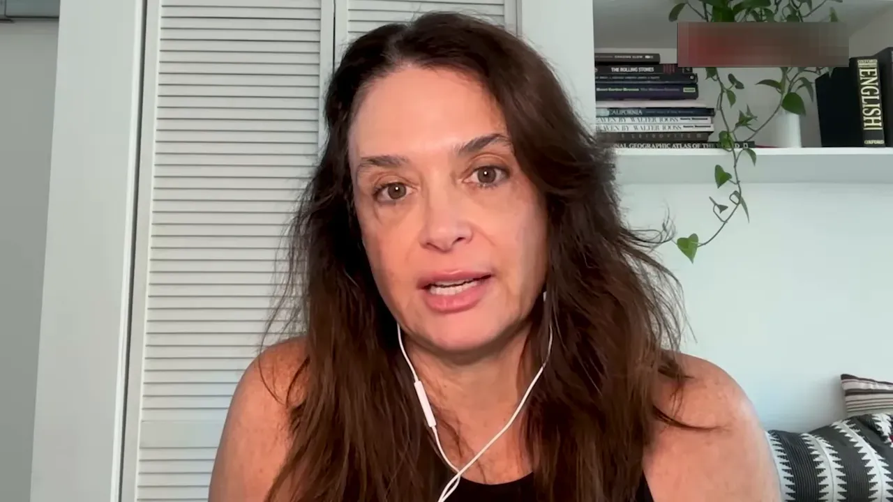 Close-up of the interview subject speaking in a well-lit room with a bookshelf and hanging plant behind, earbuds visible.