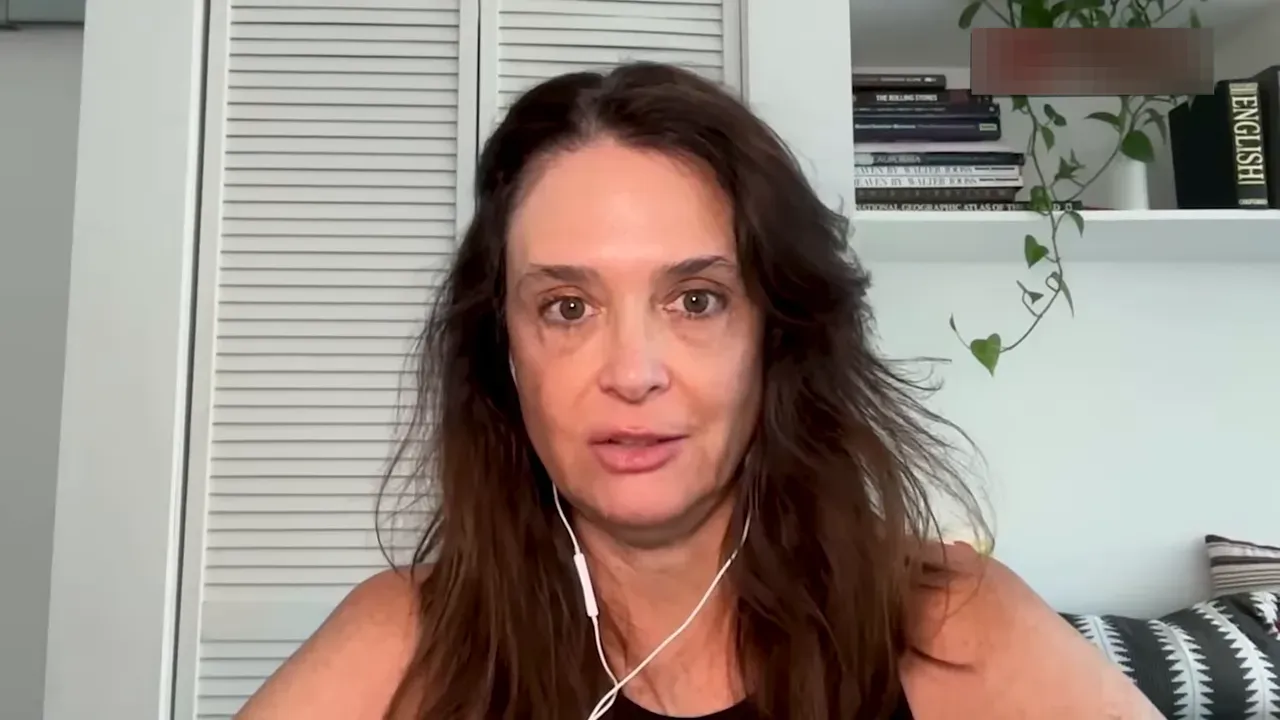 Frontal video-call portrait of an interviewee making direct eye contact with a neutral bookshelf background and a trailing plant.