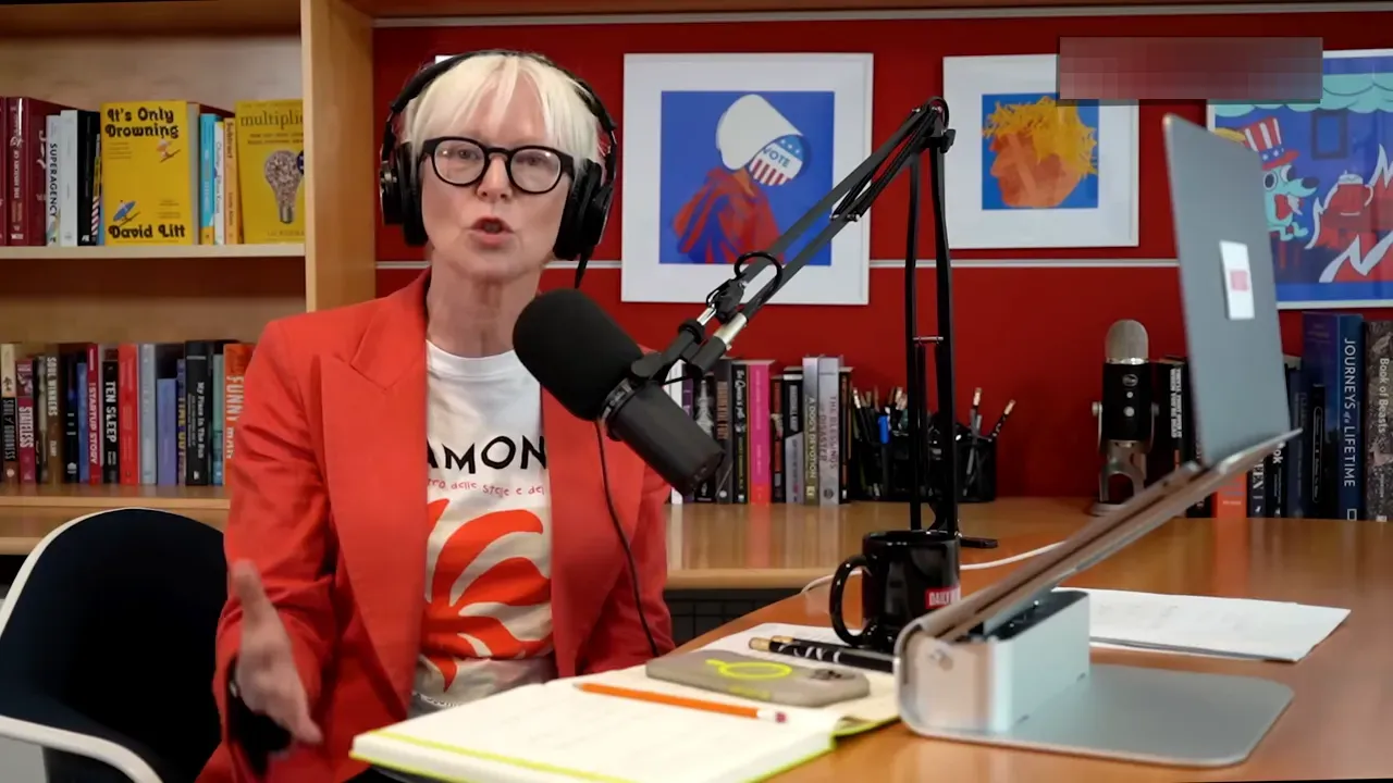 Podcast host at a studio desk wearing headphones and speaking into a microphone with books and artwork behind her.
