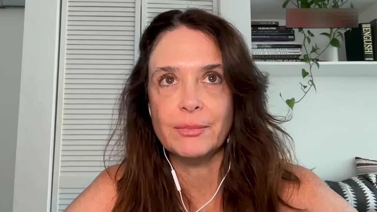 Centered close-up of interview subject looking slightly upward, earbuds visible, bookshelf and plant in background.