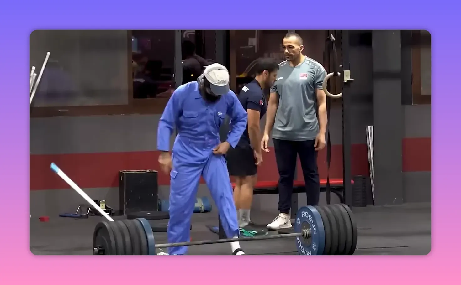 person in blue coveralls and cap standing near a heavily loaded barbell in a gym