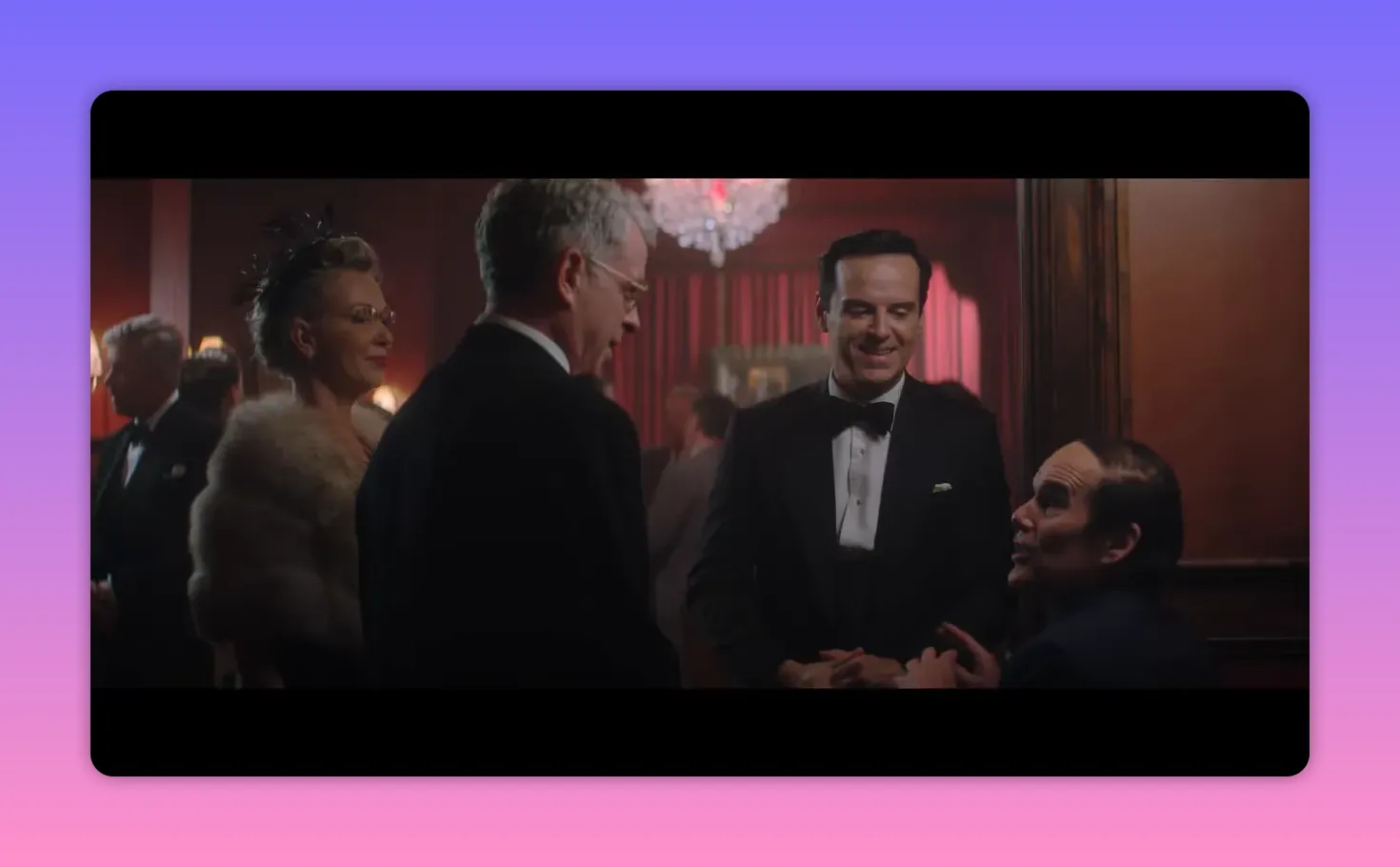Three men in formalwear talking beneath a chandelier in a crowded reception room, one man smiling in the center