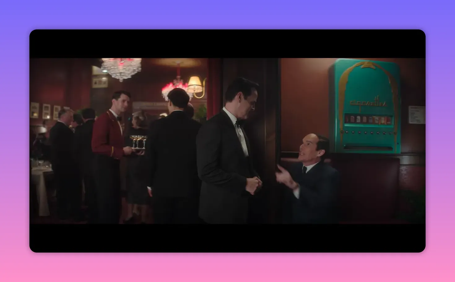 Wide shot of a packed dining room with chandeliers, a waiter carrying drinks, and two men talking near a booth
