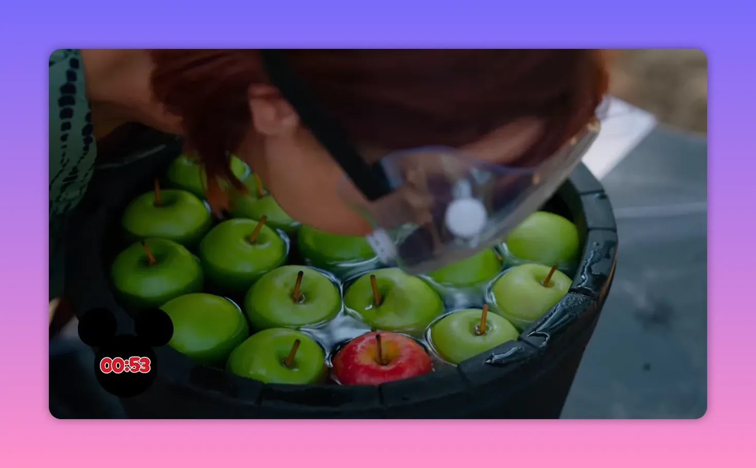 Close-up of green apples and a single red apple floating in a water tub while a contestant wearing safety goggles leans in to try and bite it.