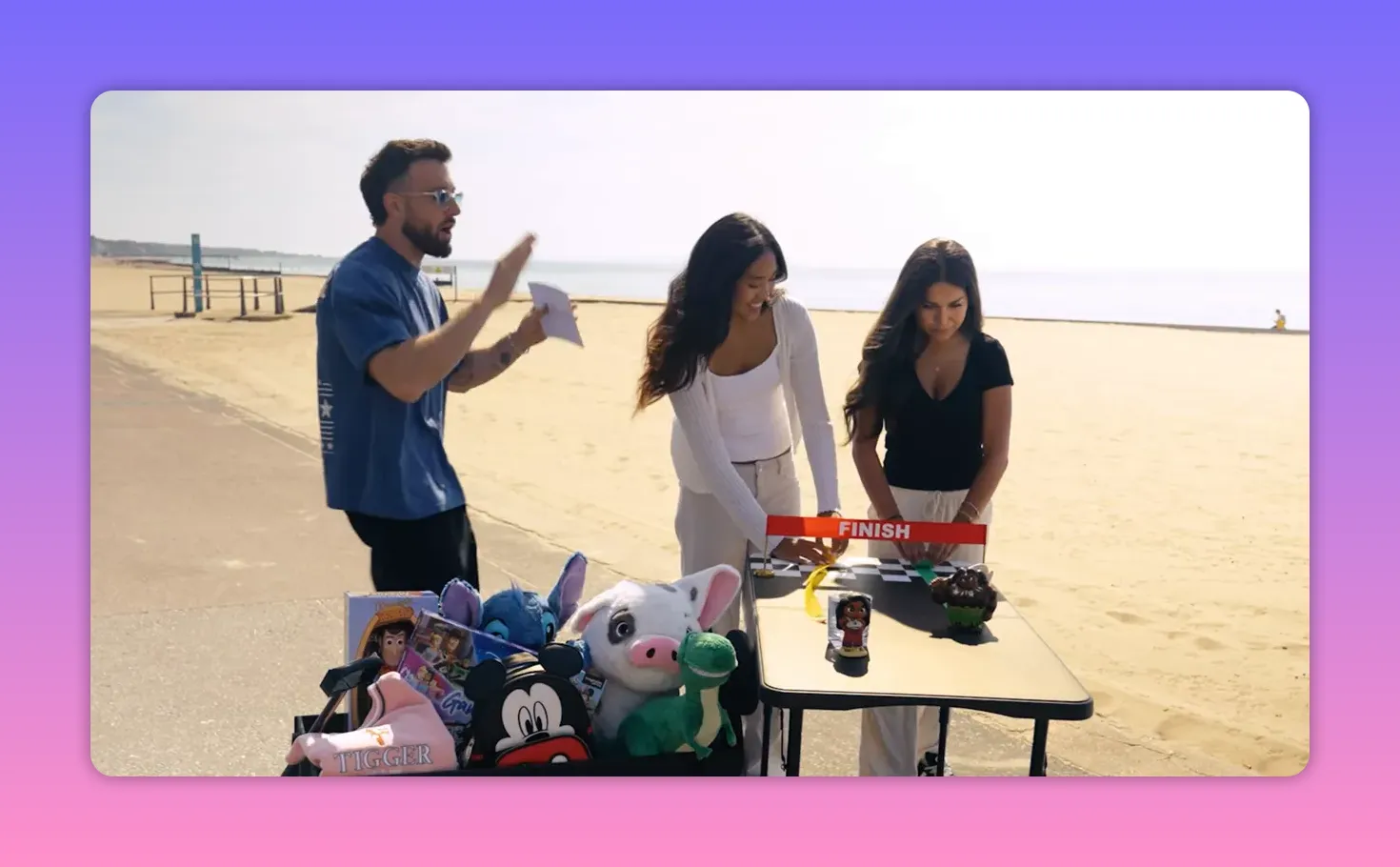 beachside game setup showing a cart full of plush prizes, a finish banner on a table, the host and two contestants