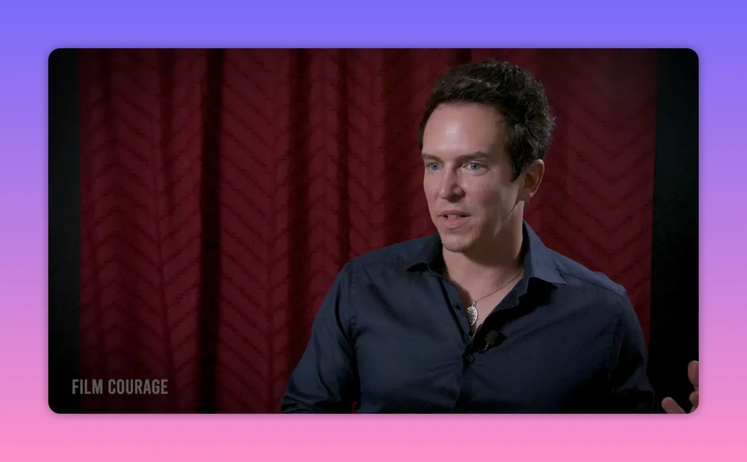 Well-lit mid-shot of an interview subject looking engaged and slightly smiling against a patterned red curtain.