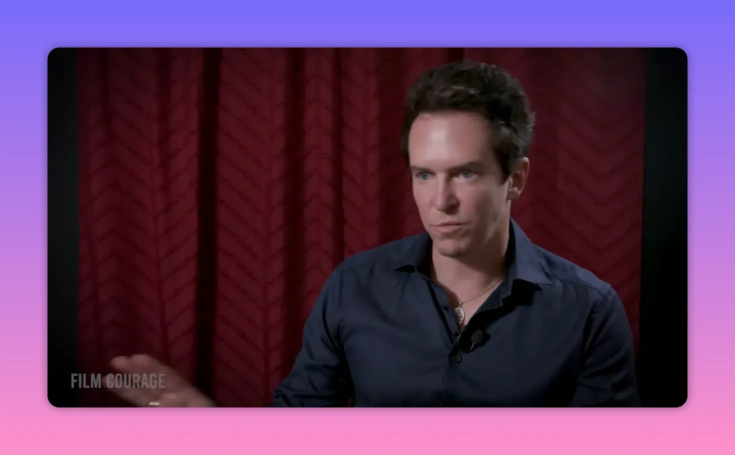 Interview shot showing a person gesturing with one hand while speaking in front of a red curtain, suggesting emphasis and instruction.