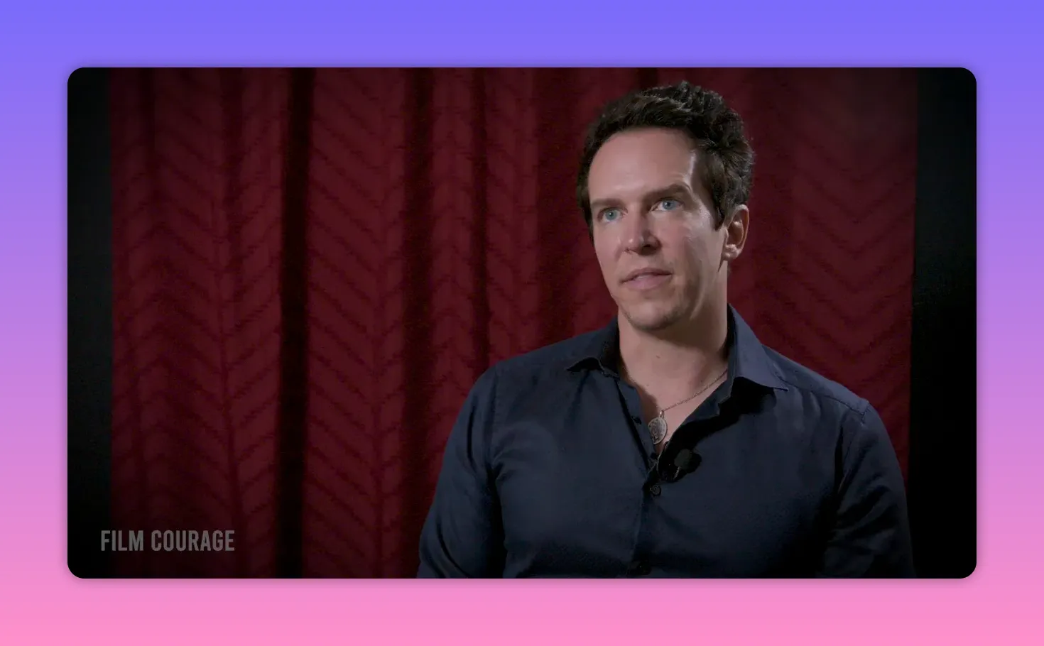 Well-lit mid-shot of an interview subject looking engaged against a patterned red curtain, wearing a dark shirt and a lavalier mic.
