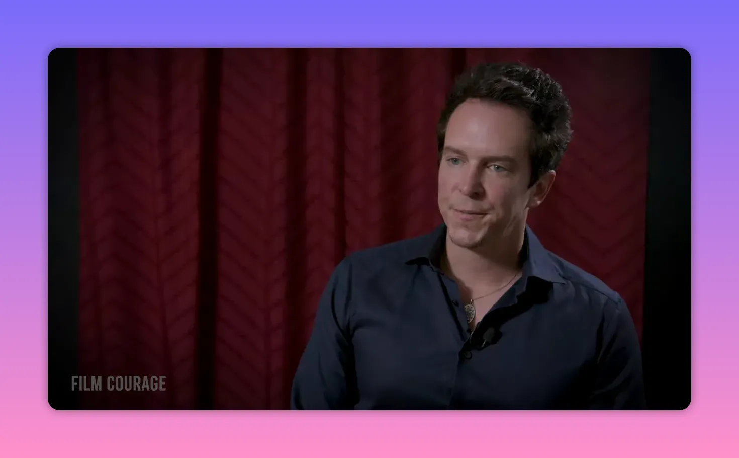 Clear, well-lit three-quarter interview shot of a filmmaker looking composed and attentive against a red curtain backdrop.