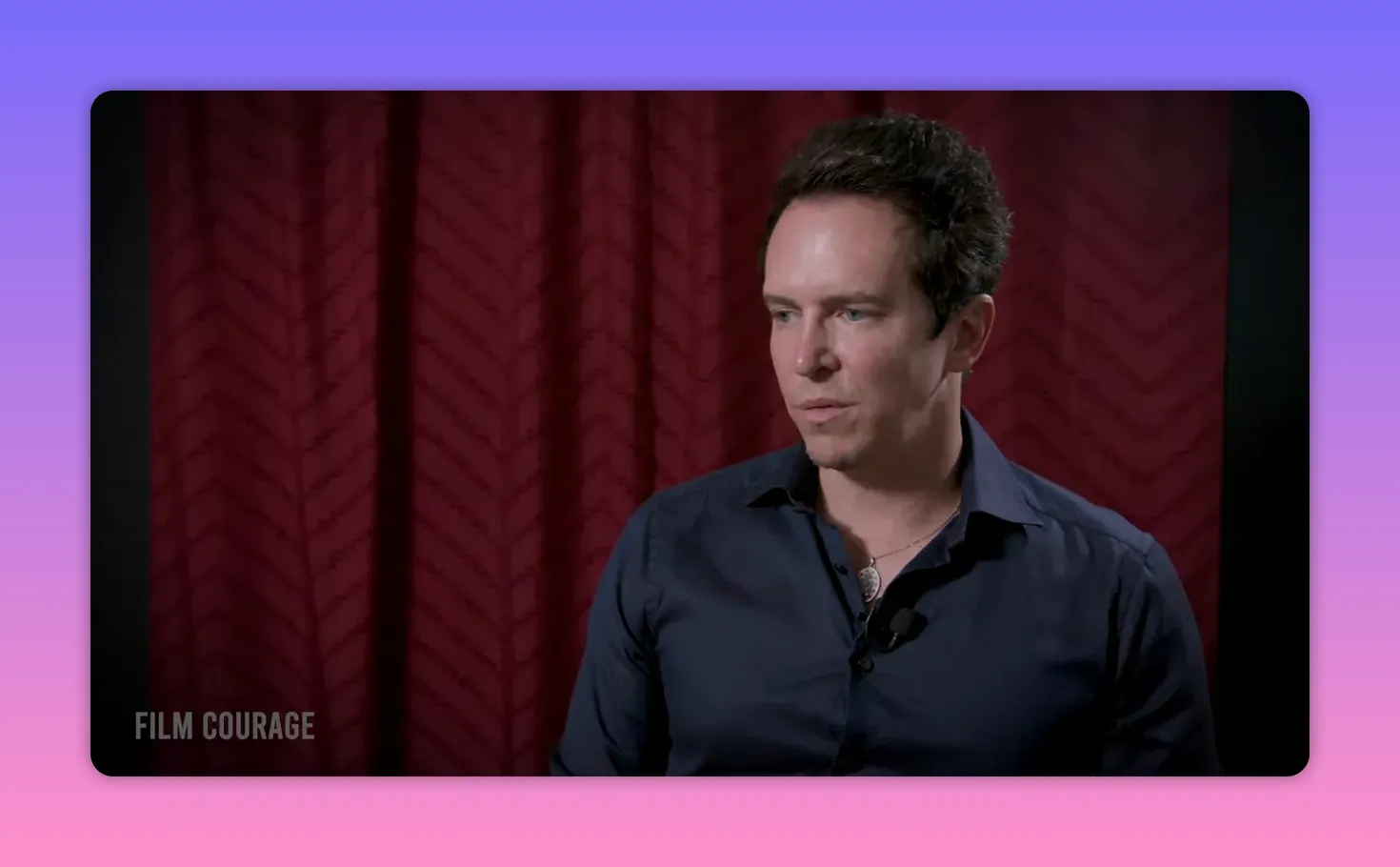 Well-composed mid-shot of a person looking thoughtful and slightly turned away, seated before a patterned red curtain.