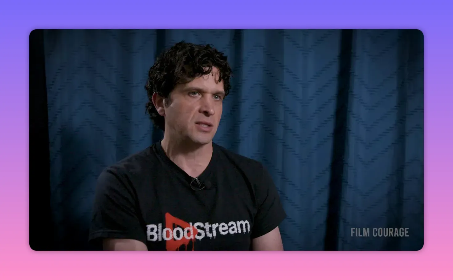 Clear mid-shot interview of a speaker wearing a BloodStream t-shirt against a blue curtain, speaking about platform business model and pricing tiers.